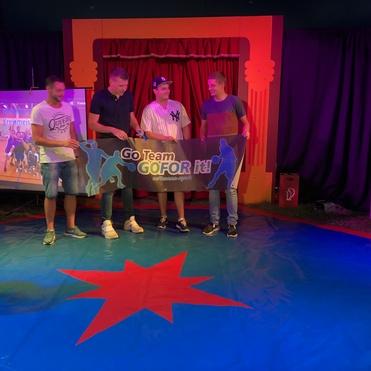 Vier Personen stehen auf einer bunten Bühne und halten ein Banner mit der Aufschrift „Go Team GOFOR it!“.