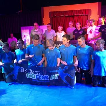 Gruppe von Jugendlichen in blauen T-Shirts posiert vor einer Bühne mit Banner „GO Team GO FOR it!“.
