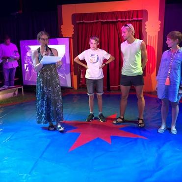 Vier Personen stehen in einem bunten Zirkuszelt, während eine Frau mit Mikrofon spricht und andere Zuschauer zuhören.