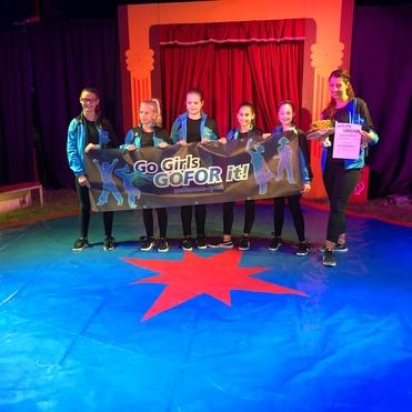 Sechs Kinder stehen auf einem Zirkusplatz und halten ein Banner mit der Aufschrift „Go Girls GO FOR it!“.