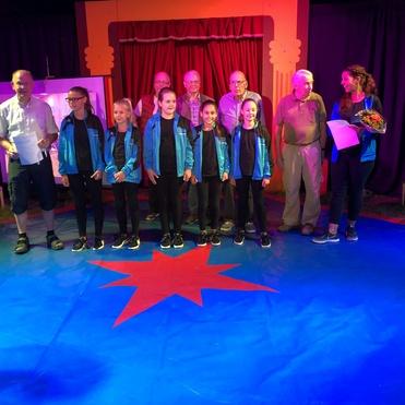 Gruppenszene auf einer Bühne mit Kindern in blauen Jacken und Erwachsenen, umgeben von buntem Vorhang und einer blauen Plane.