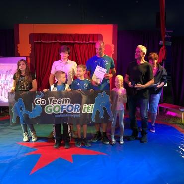 Gruppenbild von Kindern und Erwachsenen in einem Zelt, die ein Banner halten und Urkunden entgegennehmen.