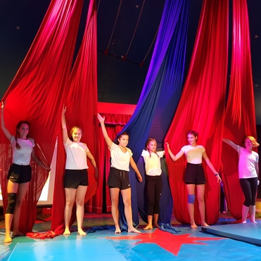 Sechs Tänzerinnen stehen auf einer Bühne in einem Zirkuszelt und halten rote und blaue Vorhänge hoch.