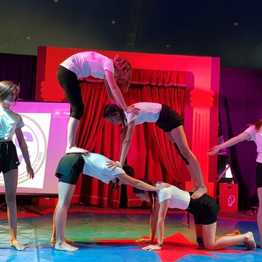 Sechs Mädchen in Sportkleidung führen eine Akrobatik-Darbietung in einem Zirkuszelt auf.