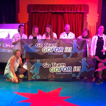 Gruppenszene auf einer Bühne mit Teilnehmern, die Banner mit „Go Team GO FOR it!“ halten. Im Hintergrund ist eine Präsentation zu sehen.