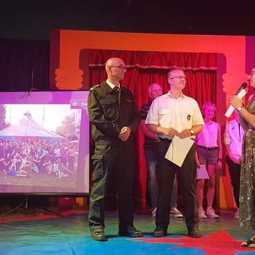 Zwei Männer in Uniform stehen auf einer Bühne, während eine Frau mit Mikrofon spricht. Im Hintergrund ein Gruppenfoto.