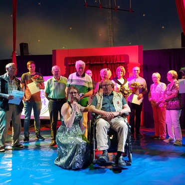 Gruppe von Menschen auf einer Bühne bei einer Ehrung, einige halten Urkunden und Blumen. Sängerin im Vordergrund.