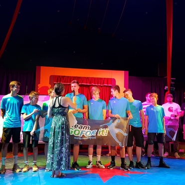 Gruppe von Jugendlichen in blauen Shirts auf einer Bühne, während eine Frau ein Mikrofon hält und mit ihnen spricht.