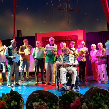 Gruppe von Menschen auf einer Bühne, einige halten Urkunden und Blumen. Ein Mann sitzt im Rollstuhl in der Mitte.