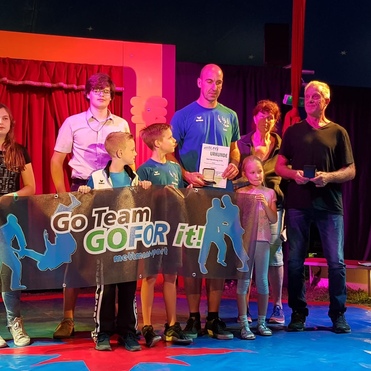 Gruppe von Personen hält ein Banner mit „Go Team GO FOR it!“, auf einer Bühne in einem Zelt, mit Urkunden und Medaillen.