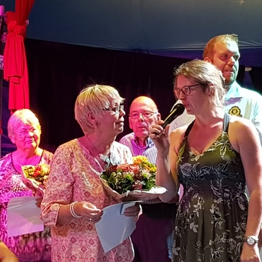 Zwei Frauen mit Blumensträußen stehen auf einer Bühne, während eine andere person am Mikrofon spricht. Hintergrund mit Zuschauern.