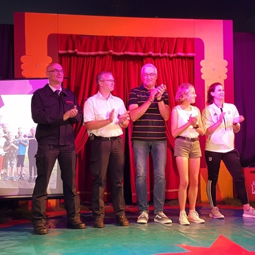 Fünf Personen applaudieren auf einer Bühne vor rotem Vorhang, im Hintergrund ein Bildschirm mit Gruppenfoto.