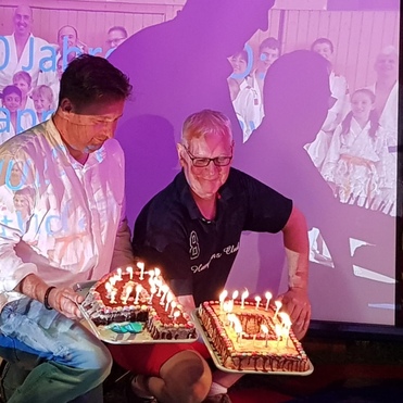 Zwei Männer halten mit Kerzen dekorierte Geburtstagstorten vor einem Projektorbild mit der Aufschrift "40 Jahre".