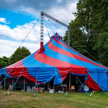 Bunt gestreifter Zirkuszelt mit blauen und roten Farben, umgeben von Bäumen und einem grünen Rasen.