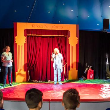 Zwei Personen stehen auf einer Bühne im Zirkuszelt, während eine dritte Person im Mittelpunkt spricht. Roter Zeltplatz.