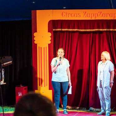 Zwei Frauen stehen auf einer Zirkusbühne vor roten Vorhängen, eine spricht ins Mikrofon, während der andere Zuschauer beobachtet.