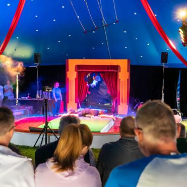 Circusaufführung mit einem Performer auf der Bühne, umgeben von rauchigen Lichtern und einem Publikum im Vordergrund.