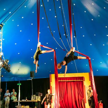 Zwei Artisten führen im Zirkuszelt akrobatische Übungen an roten Tuchbändern unter einem blauen, sternenbesäten Himmel aus.