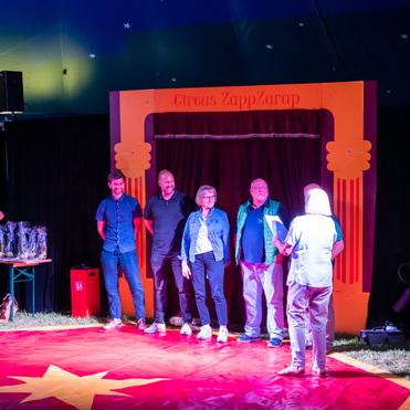 Fünf Personen stehen auf einer bunten Zirkusfläche vor einem roten Vorhang im Zelt des Circus ZappZarap.