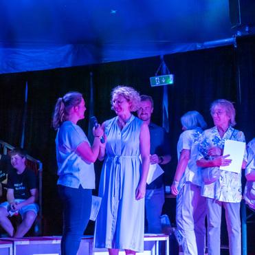 Zwei Frauen sprechen auf einer Bühne, während weitere Personen im Hintergrund stehen und lächeln. Das Licht ist blau.