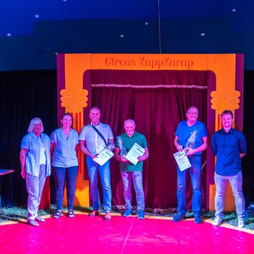 Fünf Personen stehen auf einer Bühne im Zirkus ZappZarap und halten Urkunden vor einem roten Vorhang.