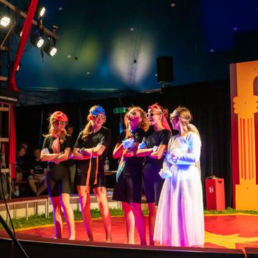 Gruppe von fünf Kindern in Kostümen auf einer Zirkusbühne, umgeben von bunten Lichtern und Kulissen.