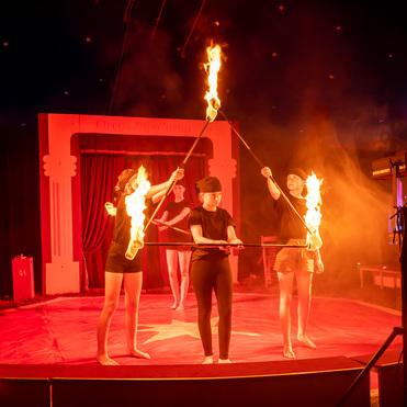 Vier Artistinnen führen eine Feuer-Performance auf einer Zirkusbühne mit roten Lichtern und Rauch durch.