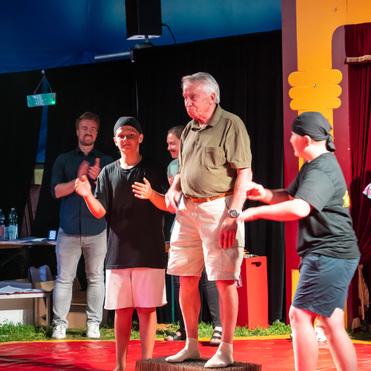 Zwei Kinder und ein älterer Mann stehen auf einer Bühne, umgeben von applaudierenden Zuschauern in einem Zirkuszelt.