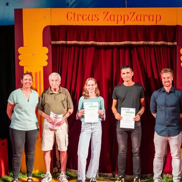 Fünf Personen stehen vor einem Vorhang im Zirkuszelt und halten Urkunden in der Hand. Hintergrund mit "Circus ZappZarap".