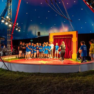 Zehn Kinder in blauen T-Shirts stehen auf einer Zirkusbühne unter einem sternenbesäten Zelt.