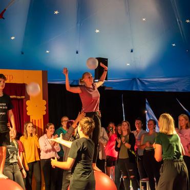 Zirkusaufführung mit Kindern, die auf großen Bällen balancieren, während das Publikum applaudiert und zuschaut.