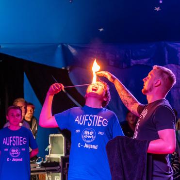 Junge mit offenem Mund hält brennenden Stock, während ein Mann ihm hilft, in einem Zirkuszelt voller Zuschauer.