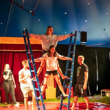 Zwei Frauen balancieren auf blauen Leitern in einem Zirkuszelt, während andere Zuschauer zuschauen.