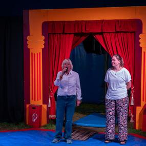 Zwei Frauen stehen auf einer blauen Fläche vor einem roten Vorhang im Zirkus, eine spricht ins Mikrofon.