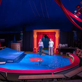 Zwei Personen stehen im Zirkus auf einer blauen Fläche mit roten Akzenten, umgeben von Zuschauerplätzen und Dekoration.