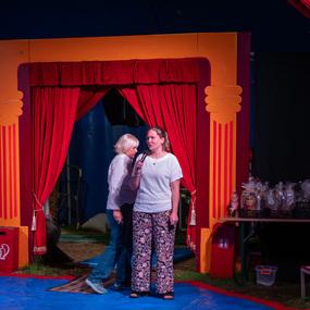 Zwei Frauen stehen auf einer Bühne im Zirkus, eine spricht ins Mikrofon, während die andere zuhört. Rote Vorhänge im Hintergrund.