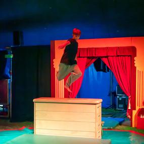 Junge springt über einen Holzblock in einem Zirkus mit roten Vorhängen und buntem Hintergrund.