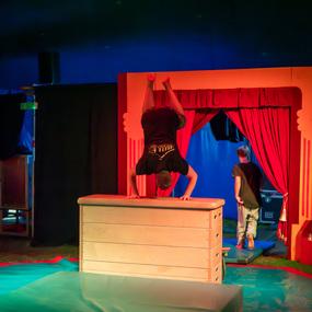 Junge macht Handstand auf einem Holzblock vor einem roten Bühnenvorhang, ein weiterer Junge geht im Hintergrund.