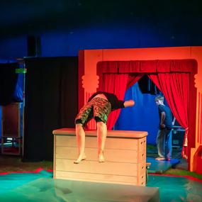 Akrobat springt über eine Holzkiste auf einer Zirkusbühne, während Zuschauer im Hintergrund beobachten.