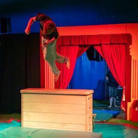 Sprung eines Jungen über einen Holztisch, im Hintergrund rote Vorhänge und eine weitere Person.