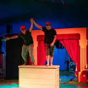 Zwei Jungen balancieren auf einer hölzernen Kiste in einem Zirkuszelt, umgeben von bunten Kulissen und Matten.