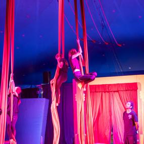 Drei Akrobatinnen üben an roten Tuchbändern in einem Zirkuszelt mit buntem, beleuchtetem Hintergrund.