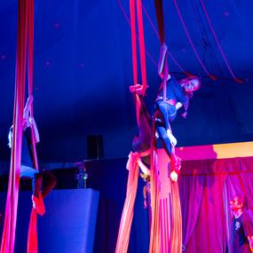 Zwei Künstlerinnen schwingen an roten Seidentüchern unter einem bunten Zelt, ein Zuschauer beobachtet die Darbietung.