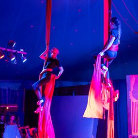 Zwei Artistinnen schwingen an roten Tuchbändern unter bunten Lichtern in einem Zirkuszelt.