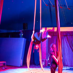 Aerial-Artist schwingt an Tüchern in einem Zirkuszelt, während zwei Personen beobachten. Farbiges Licht beleuchtet die Szene.