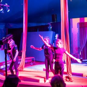 Drei Artisten in schwarzer Kleidung führen eine Aufführung im Zirkuszelt unter bunten Lichtern auf.