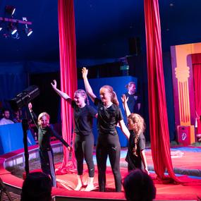 Vier junge Künstler in schwarzer Kleidung stehen im Zirkus und empfangen Applaus vor roten Vorhängen und einer farbenfrohen Kulisse.