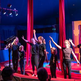 Sechs Kinder in Schwarz stehen auf einer Zirkusbühne und lächeln, während sie die Hände heben. Zuschauer klatschen.