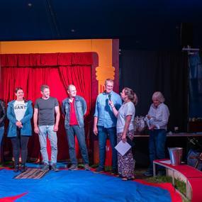 Sechs Personen stehen auf einer Zirkusbühne, während eine Frau mit Mikrofon spricht, im Hintergrund ein roter Vorhang.
