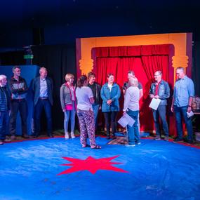 Gruppenszenen im Zelt mit mehreren Personen auf einem blauen Boden und rotem Vorhang im Hintergrund.